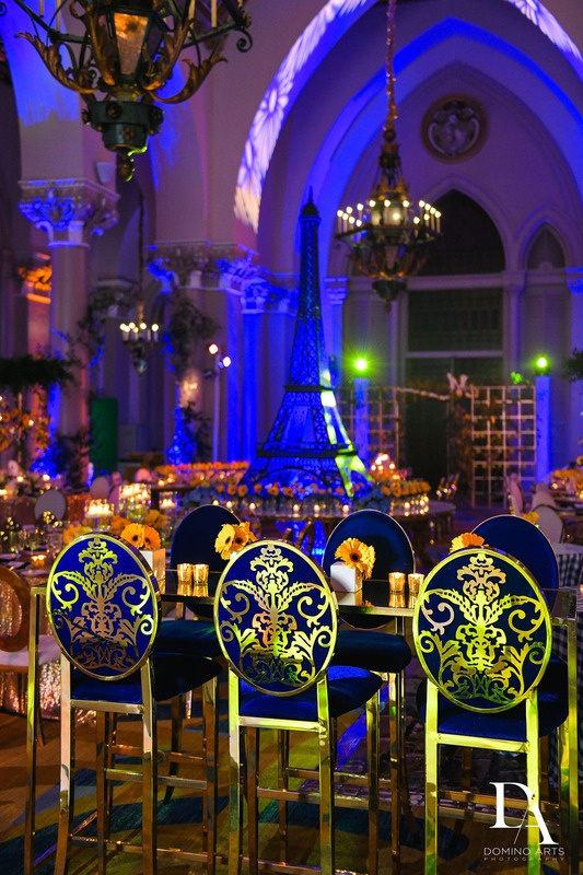 Elegant event venue decorated with a miniature Eiffel Tower centerpiece, illuminated blue projections, and sunflower-themed table settings with candles, chandeliers, and Gothic architecture in the background.