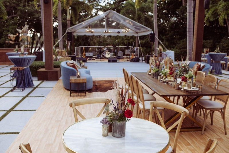 Elegant outdoor event space with a wooden dining table decorated with floral centerpieces, surrounded by wooden chairs, a separate round table with a blue cloth, a cozy lounge area with blue and gray seating, and a stage setup under a clear tent for 