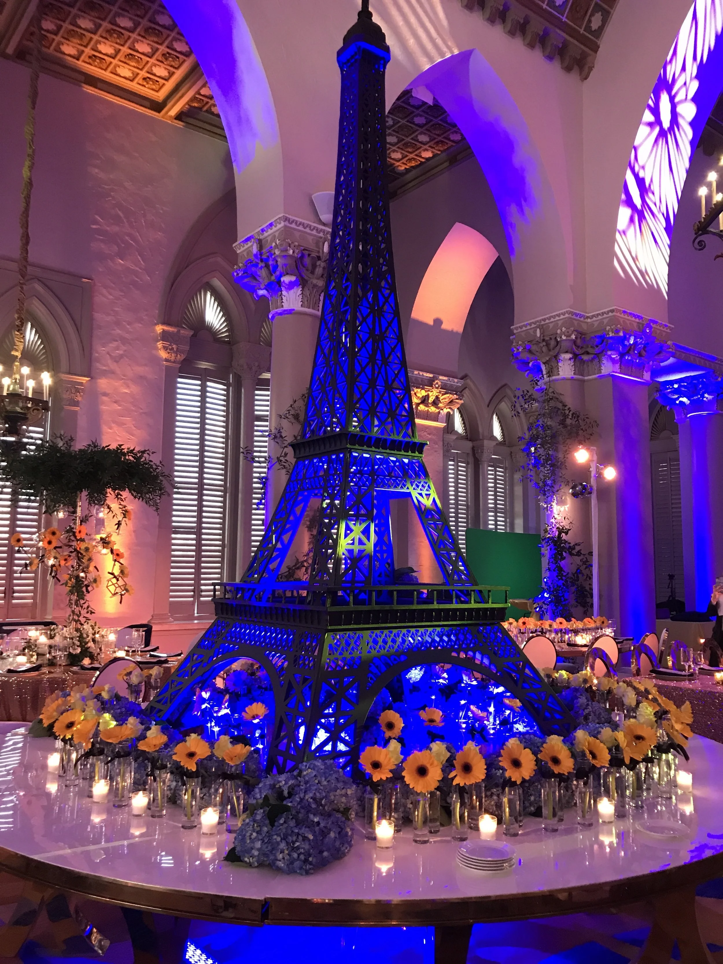 A decorative event setup featuring a large model of the Eiffel Tower illuminated with blue lighting, surrounded by a border of yellow and purple flowers, candles, and elegant tables with place settings, in a grand indoor space with tall windows and c