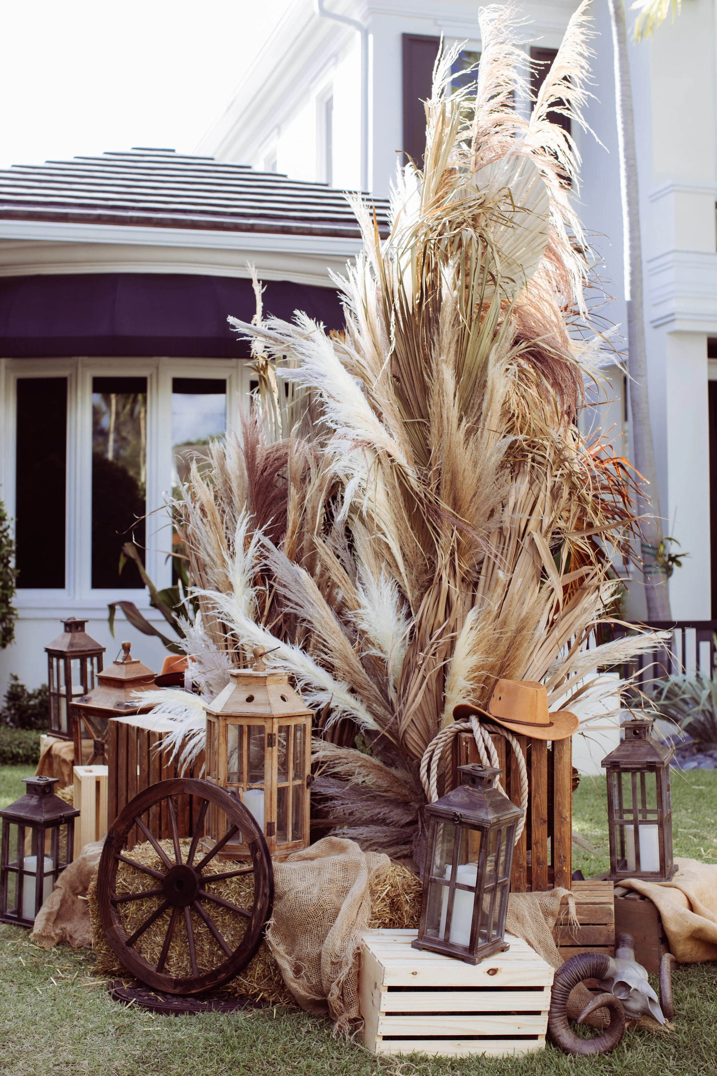 Rustic outdoor display with dried pampas grass, lanterns, wooden crates, a wagon wheel, and a cowboy hat in front of a white house with a porch and brown roof.