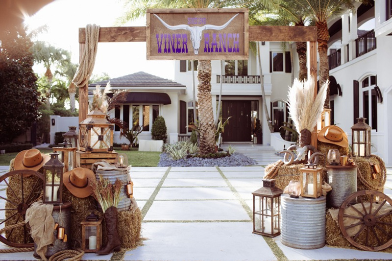Rustic outdoor wedding or event setup with hay bales, lanterns, hats, and dried plants, in front of a white house with palm trees and a sign that reads 'Viner Ranch' with a longhorn skull graphic.