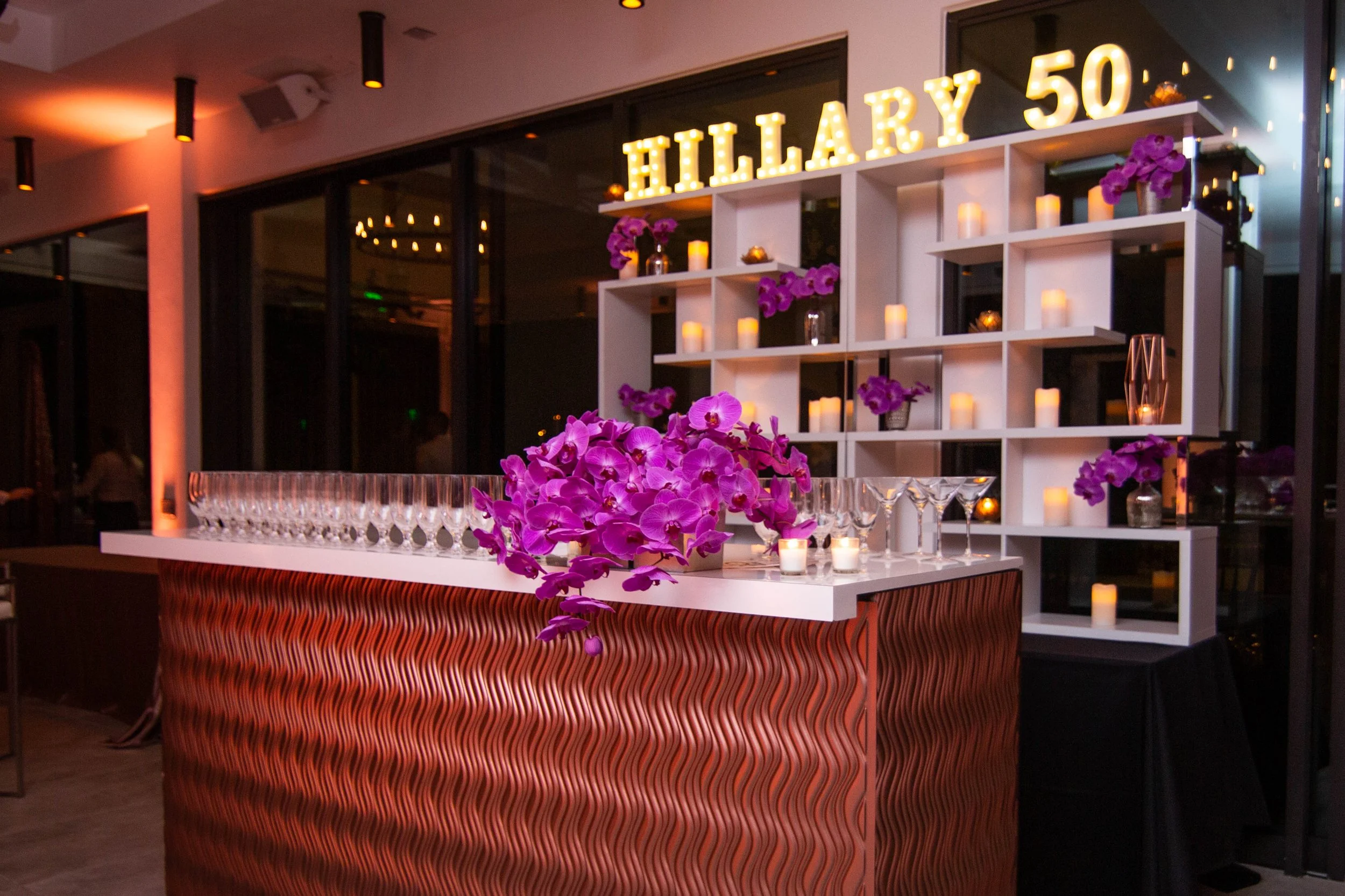 Decorative display for a 50th birthday celebration with illuminated sign reading 'Hillary 50', purple orchids, candles, and glassware on a colored table, set inside a venue with large windows.