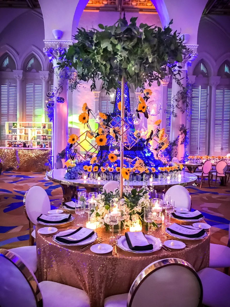 A decorated banquet table with sunflower centerpieces, candles, and place settings, within an ornate, brightly lit venue with arched windows and purple and blue lighting.