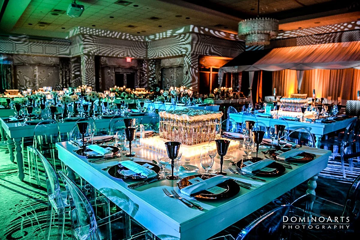 Elegant banquet hall decorated for a formal event with tables set with black and metallic dinnerware, floral centerpieces, and colorful ambient lighting.