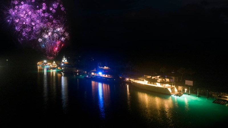 Fireworks exploding above boats docked at a marina at night, with colorful reflections on the water.