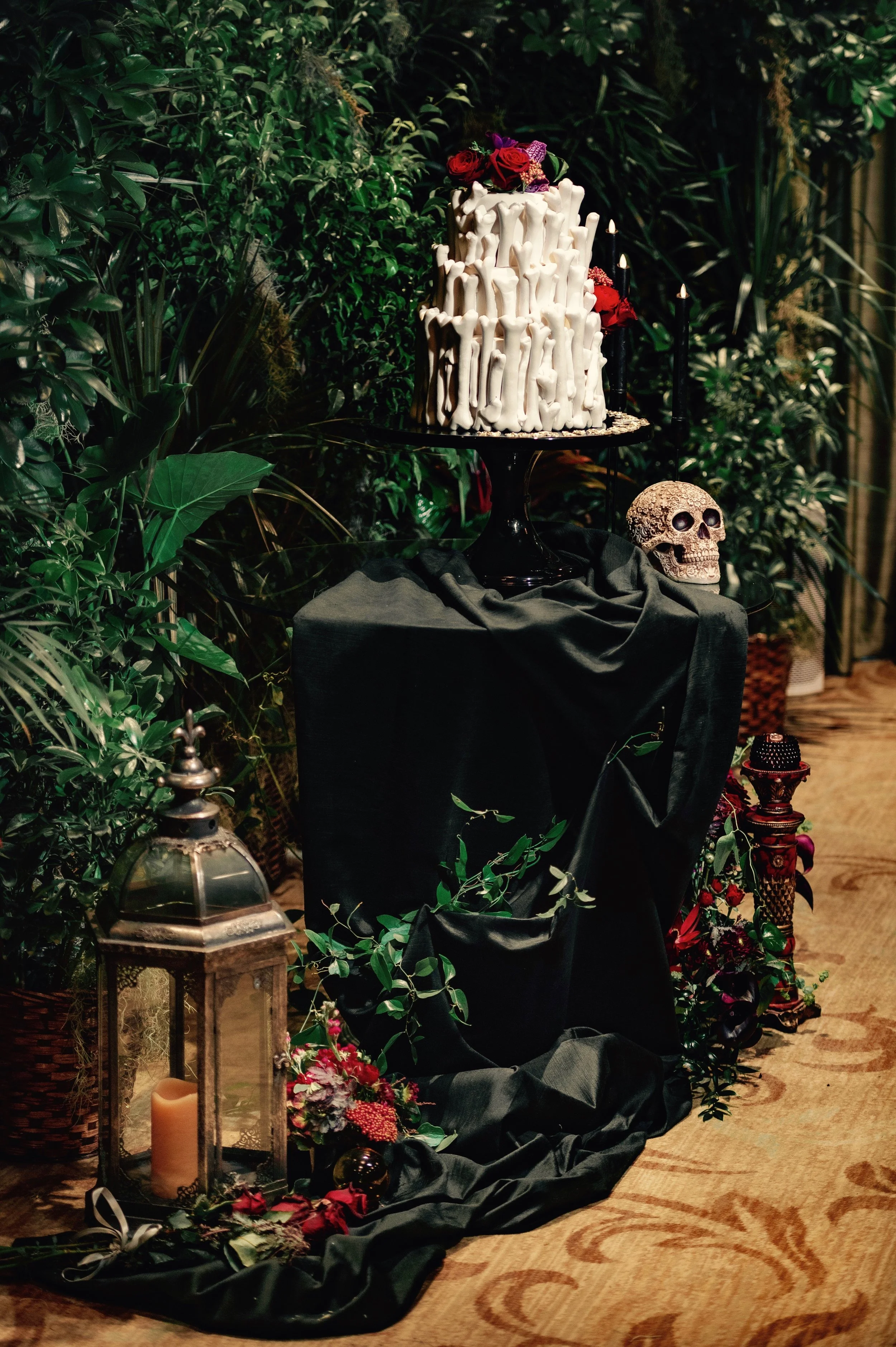 A Gothic styled display with a tall white cake decorated with bones and flowers, surrounded by dark candles, skull, candles, lantern, and greenery, on black fabric on a patterned wooden floor.
