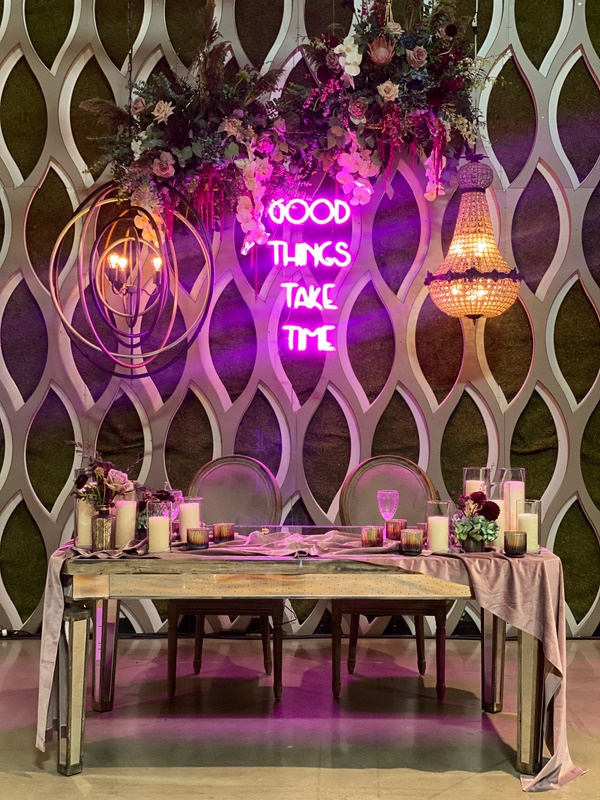 Decorative banquet table setup with candles, flowers, and pink tablecloths in front of a geometric wall. Neon sign reads 'Good Things Take Time' with floral arrangements, hanging chandeliers, and modern lighting fixtures.