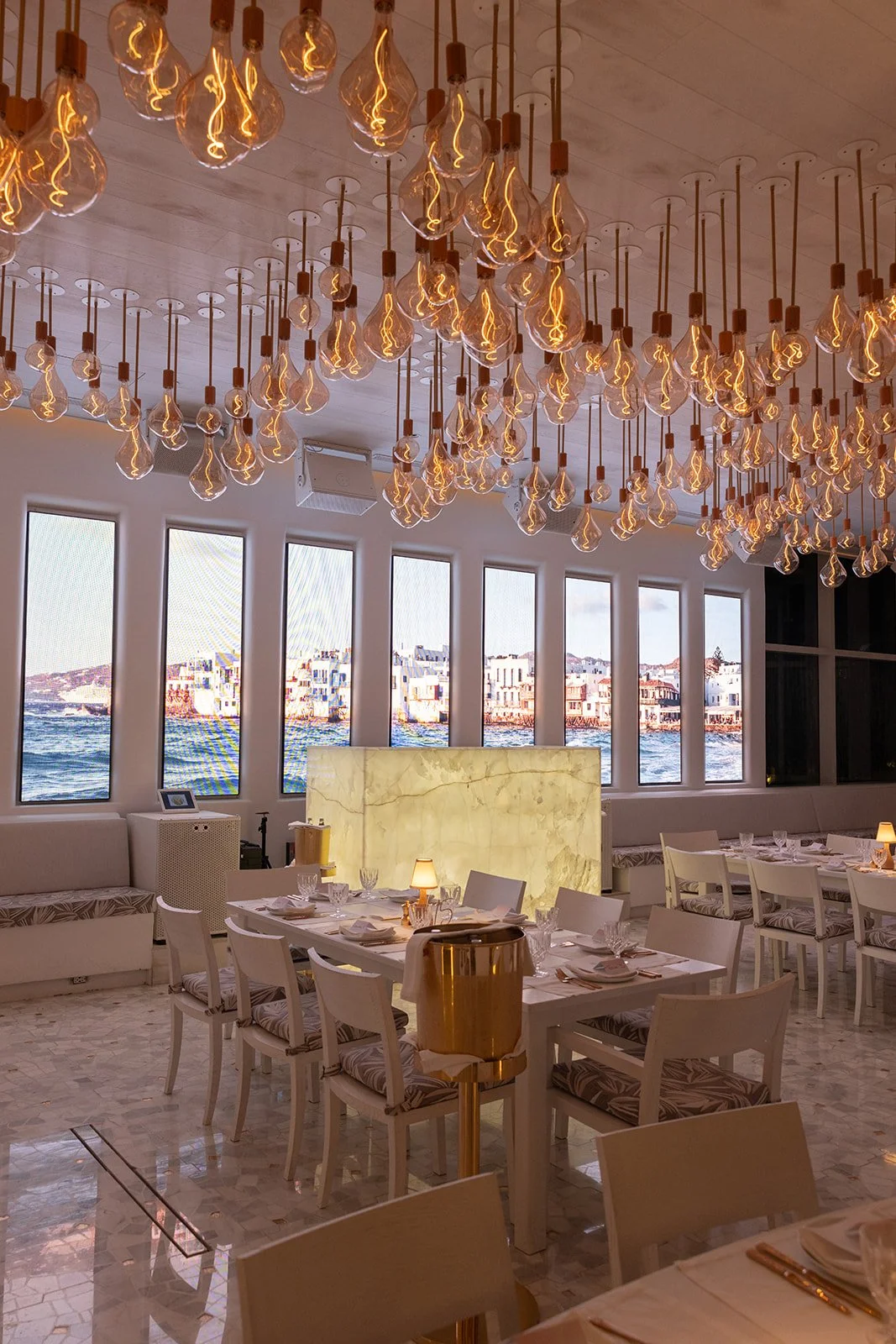 Interior of a stylish restaurant with large windows overlooking water, decorated with hanging Edison-style light bulbs on the ceiling, white chairs, and tables set for dining.