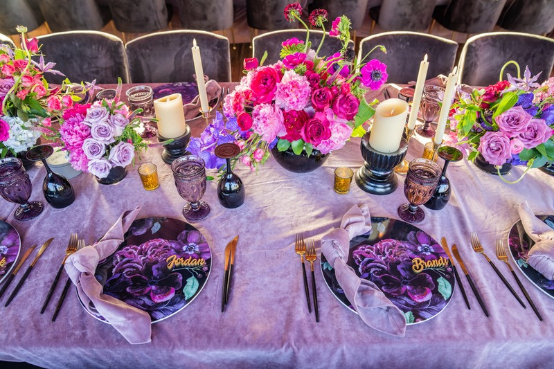 A decorated dining table with purple and pink floral centerpieces, candles, gold and purple accents, black and gold cutlery, and personalized plates with purple floral designs and gold names.
