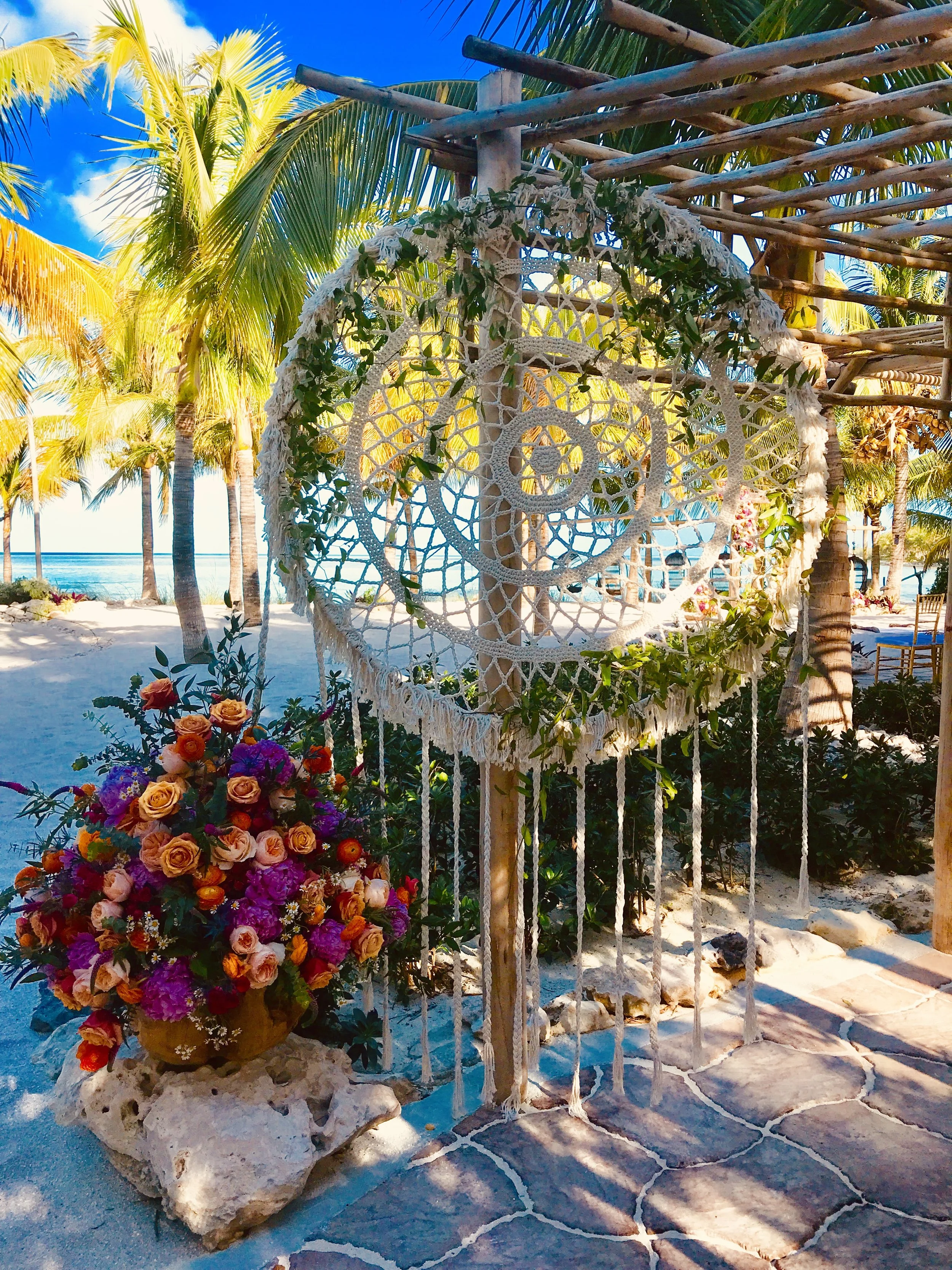Beachside scene with palm trees, a decorative hanging dreamcatcher, and a flower arrangement on a stone base in the sand, under a clear blue sky.