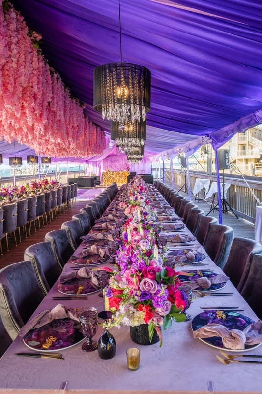 Long banquet table decorated with floral centerpieces, set for a formal event under purple draped ceiling with hanging chandeliers and pink floral decor.