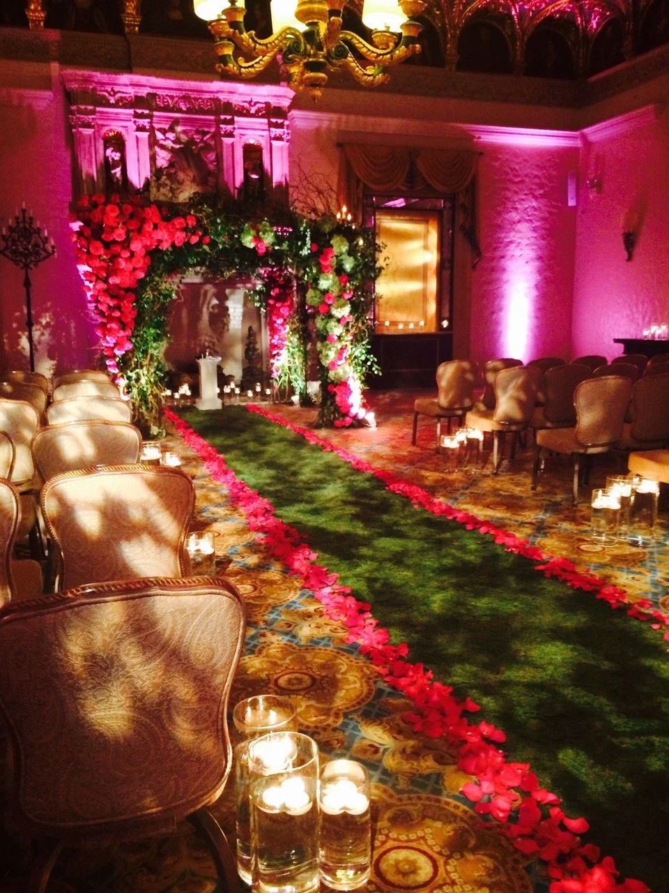 Elegant wedding ceremony decorated with flowers, greenery, and candles, set in a grand room with velvet chairs, a green carpet lined with red rose petals, and pink uplighting.