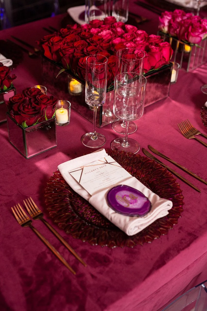 Elegant dinner table decorated with pink and red roses, glassware, candles, and a menu placed on a white napkin on a purple placemat.