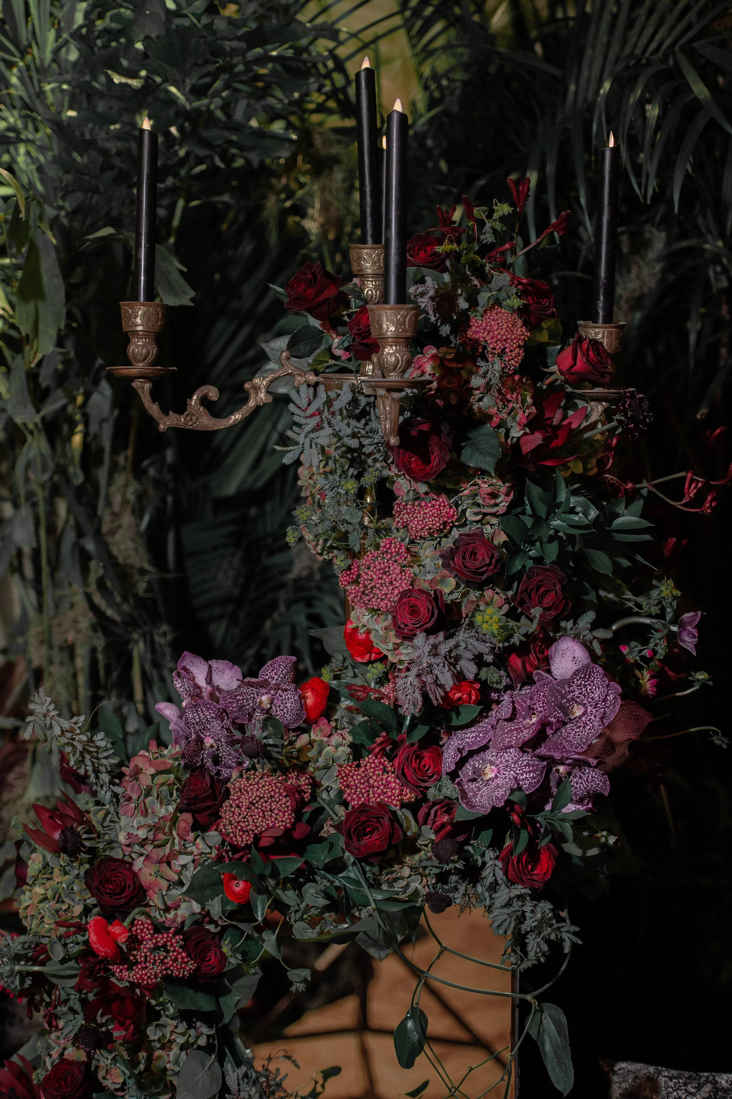 A floral arrangement with roses, orchids, and greenery on a decorative black and gold candelabrum with black candles. The background is filled with lush green plants.