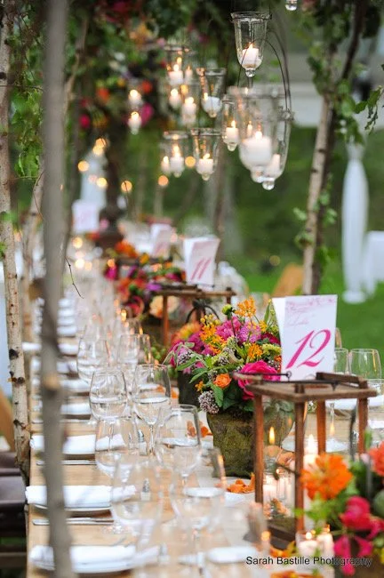 Outdoor event table decorated with colorful flowers, lanterns, and candles, with place settings and numbered table cards.
