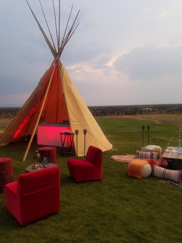 Outdoor gathering setup featuring a large beige tipi with wooden poles, in a vast green field during dusk, with red chairs, small tables, colorful cushions, and decorative lighting.