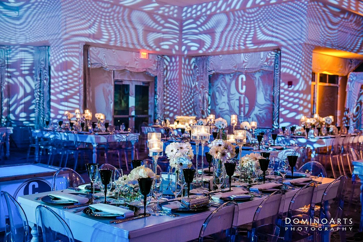 Decorated banquet hall with long tables, elegant white and black table settings, and floral centerpieces illuminated by candlelight and ambient lighting.