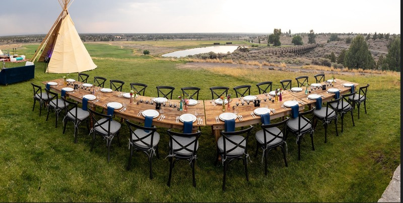 Outdoor dining table set for a large gathering with chairs, plates, napkins, and utensils, with a teepee and landscape view in the background.