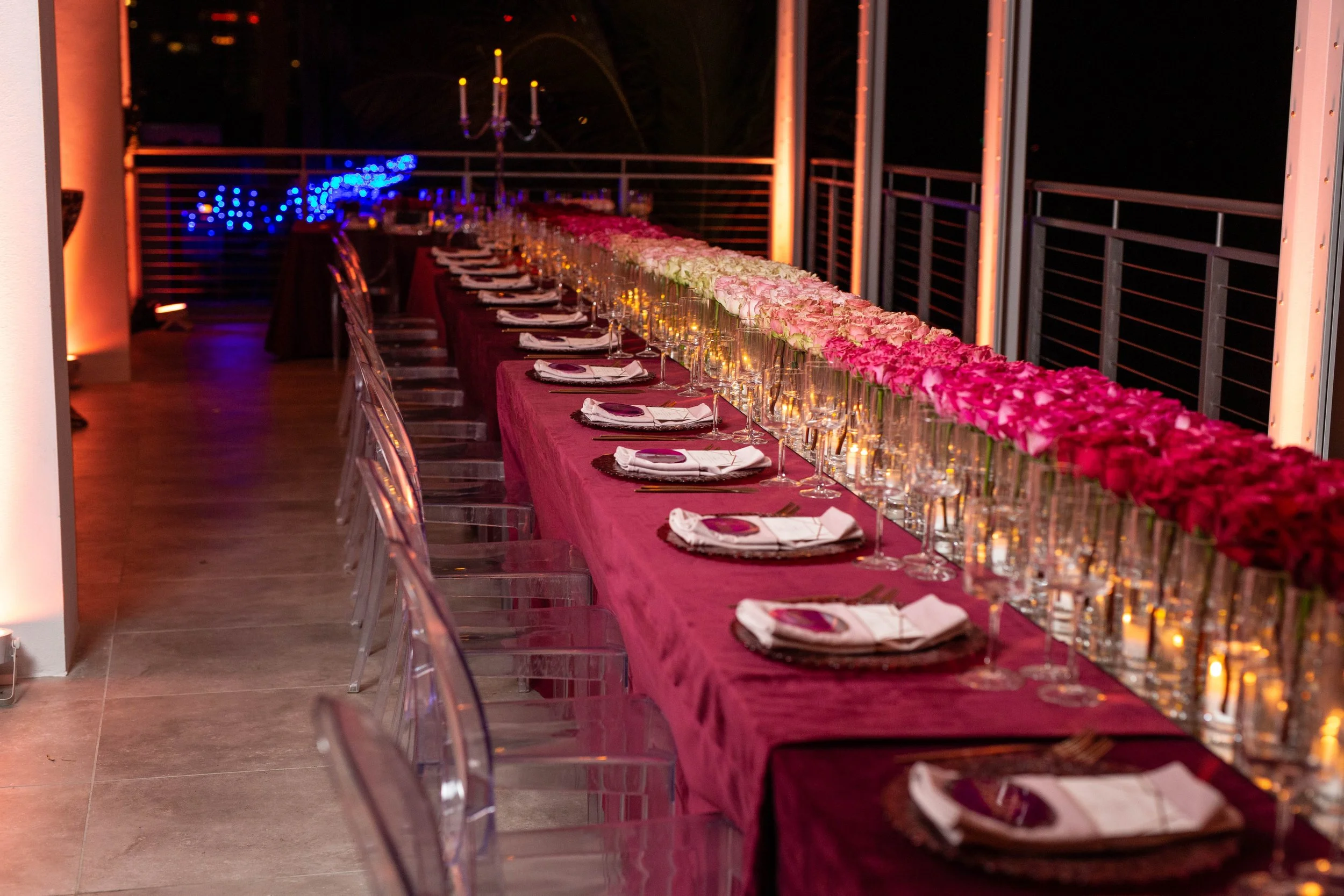 A long dining table decorated with pink floral arrangements, set with white napkins, wine glasses, and plates, in an outdoor space at night with ambient lighting and a chandelier overhead.