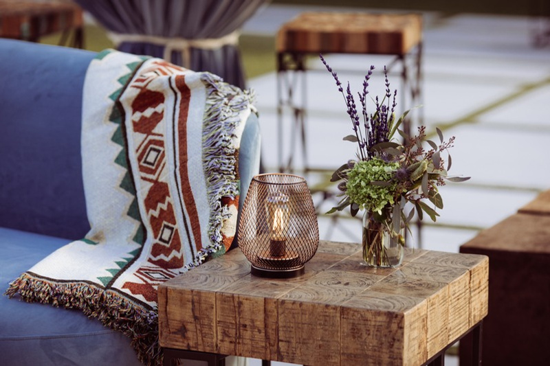 A wooden side table with a candle lantern and a vase of purple and green flowers next to a blue sofa with a patterned throw blanket. There are outdoor chairs and a patio in the background.