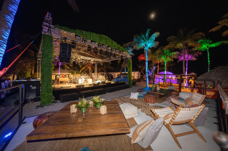 Outdoor nightclub or event space at night with a stage with musical instruments, colorful illuminated palm trees, and seating area with cushions and a wooden table.