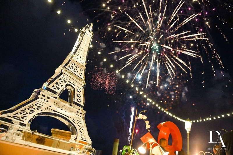 Fireworks exploding in the night sky above a large Eiffel Tower model on a rooftop or event space, with decorative lights and large red and white ornaments in the foreground.