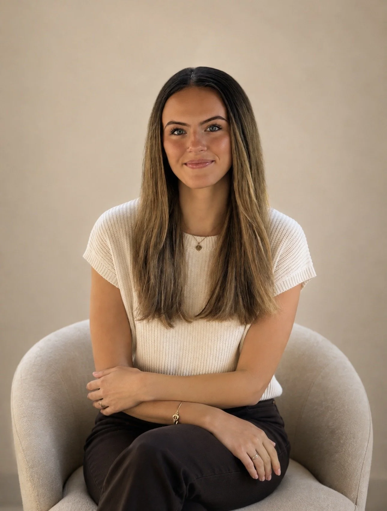 Young woman with long brown hair sitting on a beige armchair against a neutral background, smiling at the camera.