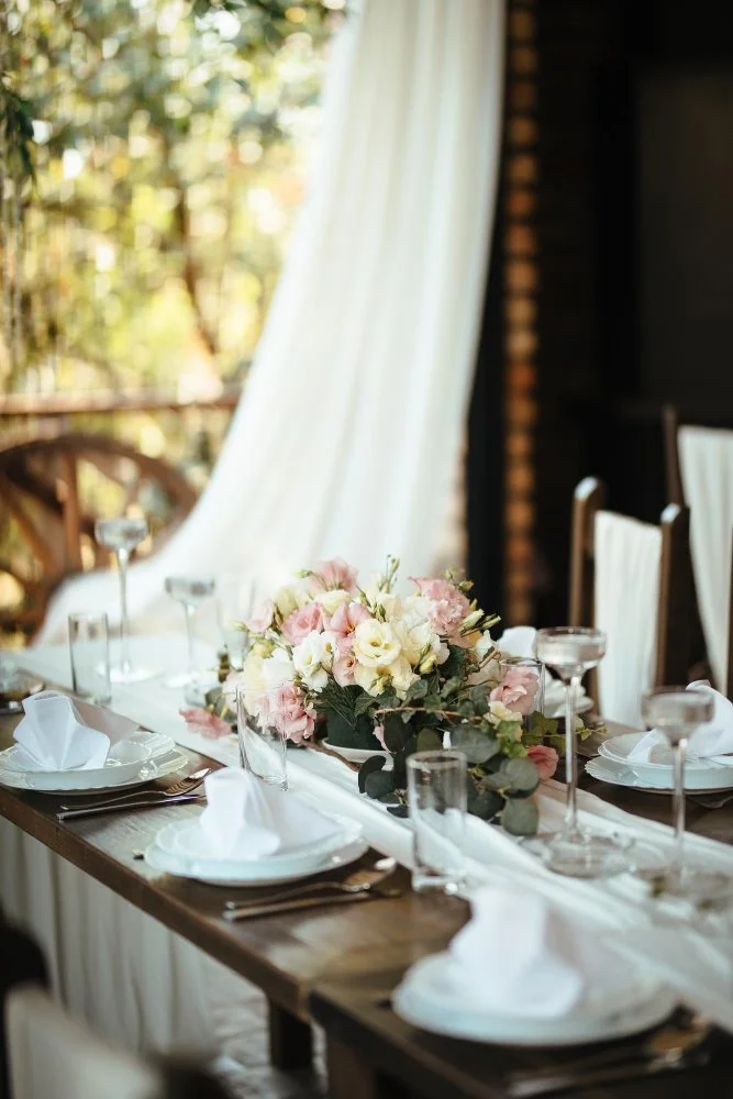Elegant table setting with white plates, napkins, glassware, and a floral centerpiece of pink and white flowers, set in a cozy indoor space with natural light and sheer curtains.