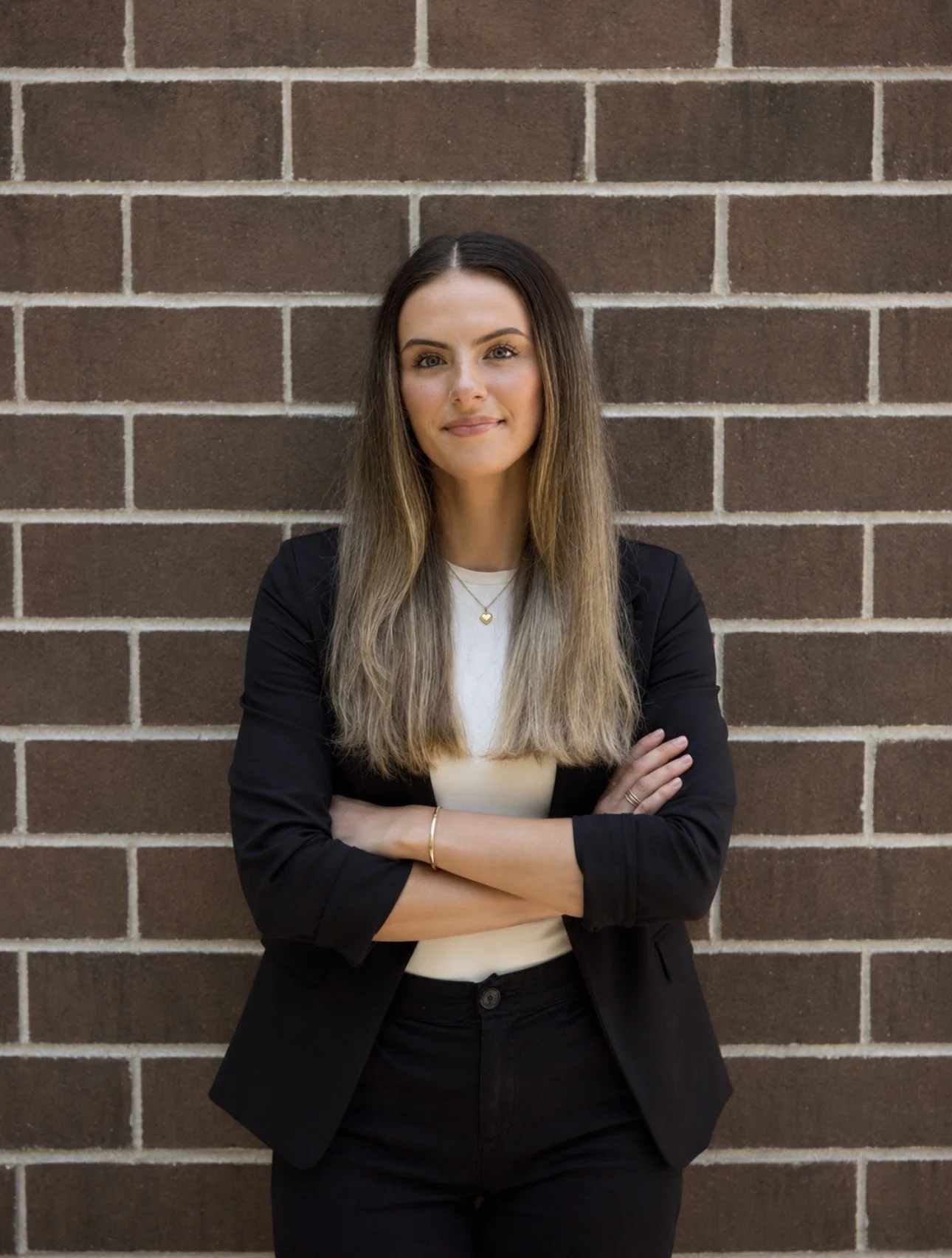 A woman with long, light brown hair, wearing a black blazer over a white top, standing with arms crossed in front of a brick wall.