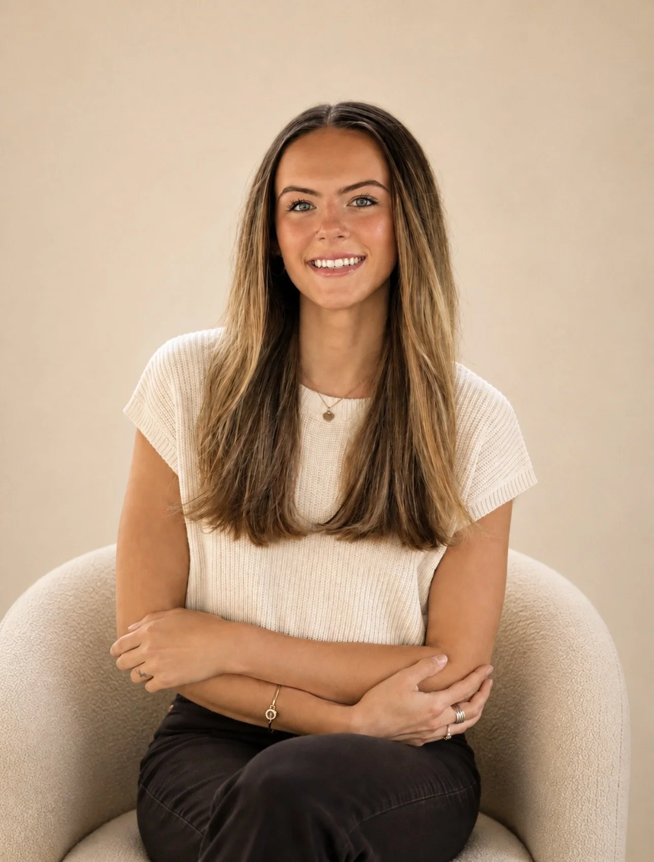 A young woman with long, wavy brown hair sitting on a beige armchair, smiling at the camera, wearing a cream short-sleeve top and black pants.