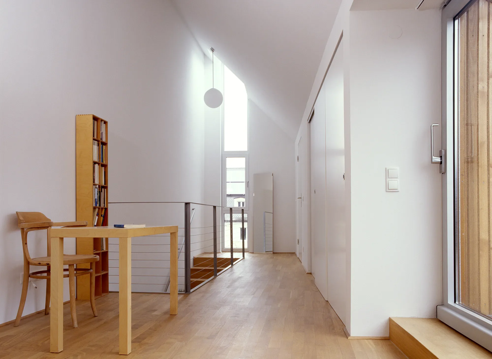 Innenaufnahme eines modernen Hauses mit Holzfußboden, weißen Wänden und großen Fenstern. Es gibt einen kleinen Holztisch mit Stühlen und eine Bücherregel. Das Licht fällt durch die Fenster, und es gibt eine Deckenleuchte.