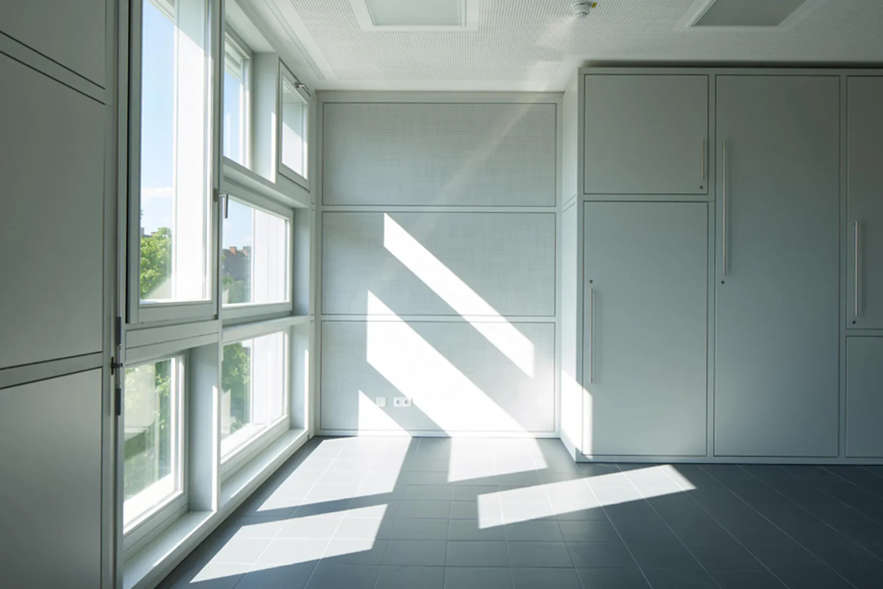 Ein heller, modern gestalteter Raum mit großen Fenstern, durch die Sonnenlicht einfällt. Weißer Wandanstrich mit Bildschirmen und grauen Fliesen auf dem Boden.