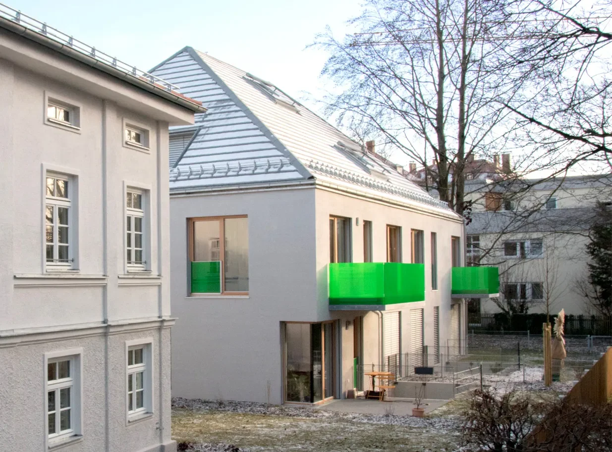 Neues Mehrfamilienhaus mit weißen Wänden und grünen Balkonen, im Hintergrund Bäume und ältere Häuser