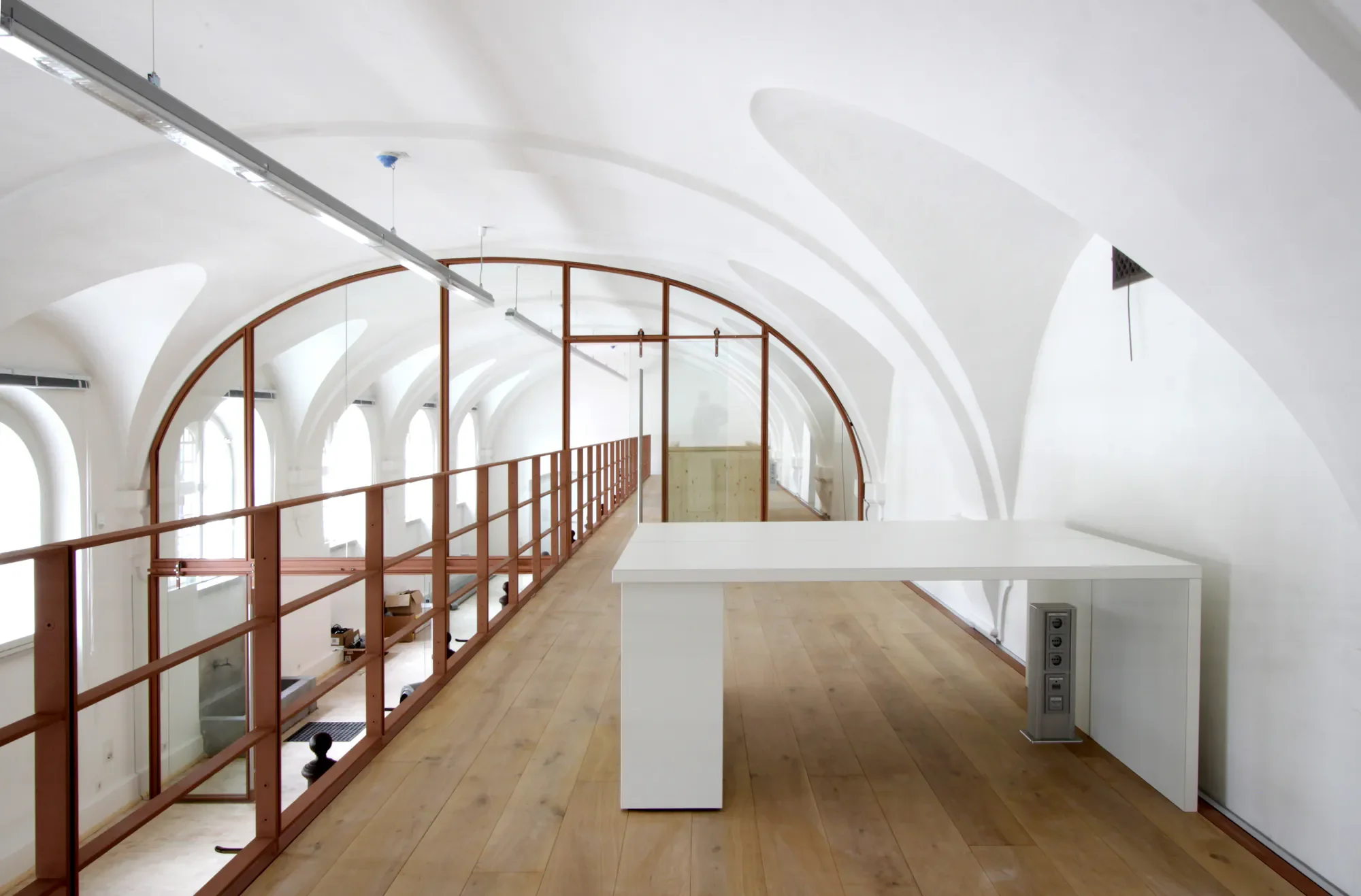 Innenansicht eines weißen, gewölbten Raumes mit Holzboden, einer Balustrade aus Holz, einer weißen Theke, mehreren Stromanschlüssen an einer weißen Wand und großen Fenstern. Der Raum ist leer, es sieht aus, als befindet sich im Aufbau.