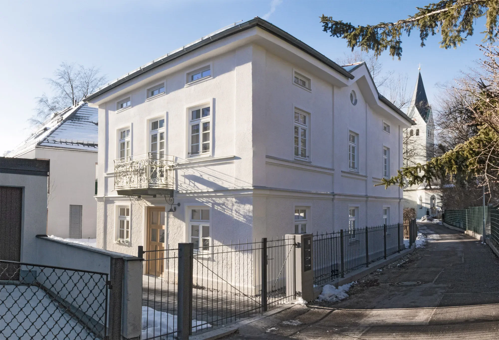 Ein weißes, drei-geschossiges Haus mit mehreren Fenstern und einem kleinen Balkon. Im Hintergrund ist eine Kirchturm mit spitzem Dach. Es sind Schneereste auf dem Dach und am Boden sichtbar, da es Winter ist.