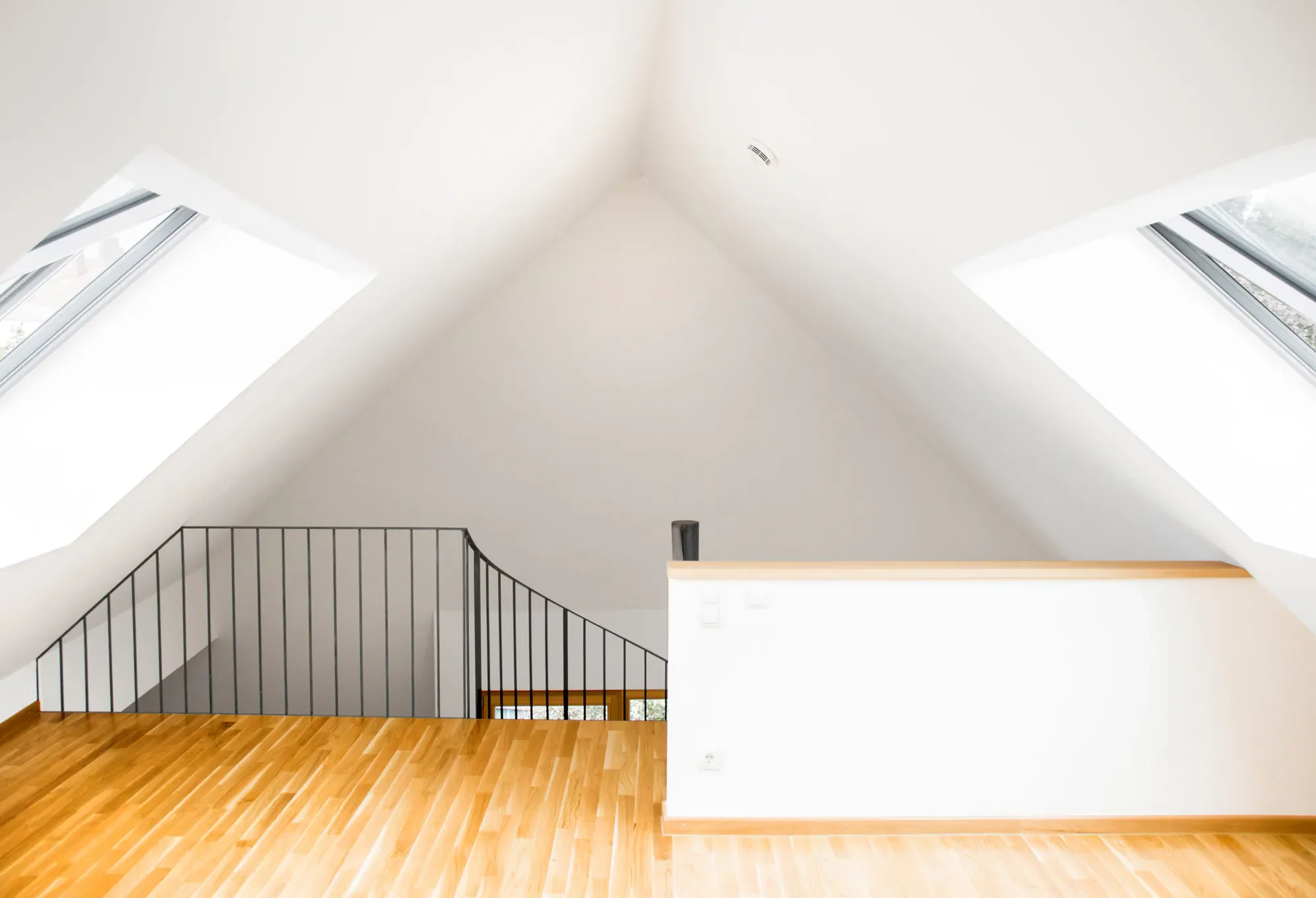 Ein heller, moderner Raum mit weißen Wänden und einem hohen, dreieckigen Dach mit großen Dachfenstern. Es gibt eine Holztreppe mit schwarzen Geländern und einen Holzfußboden.