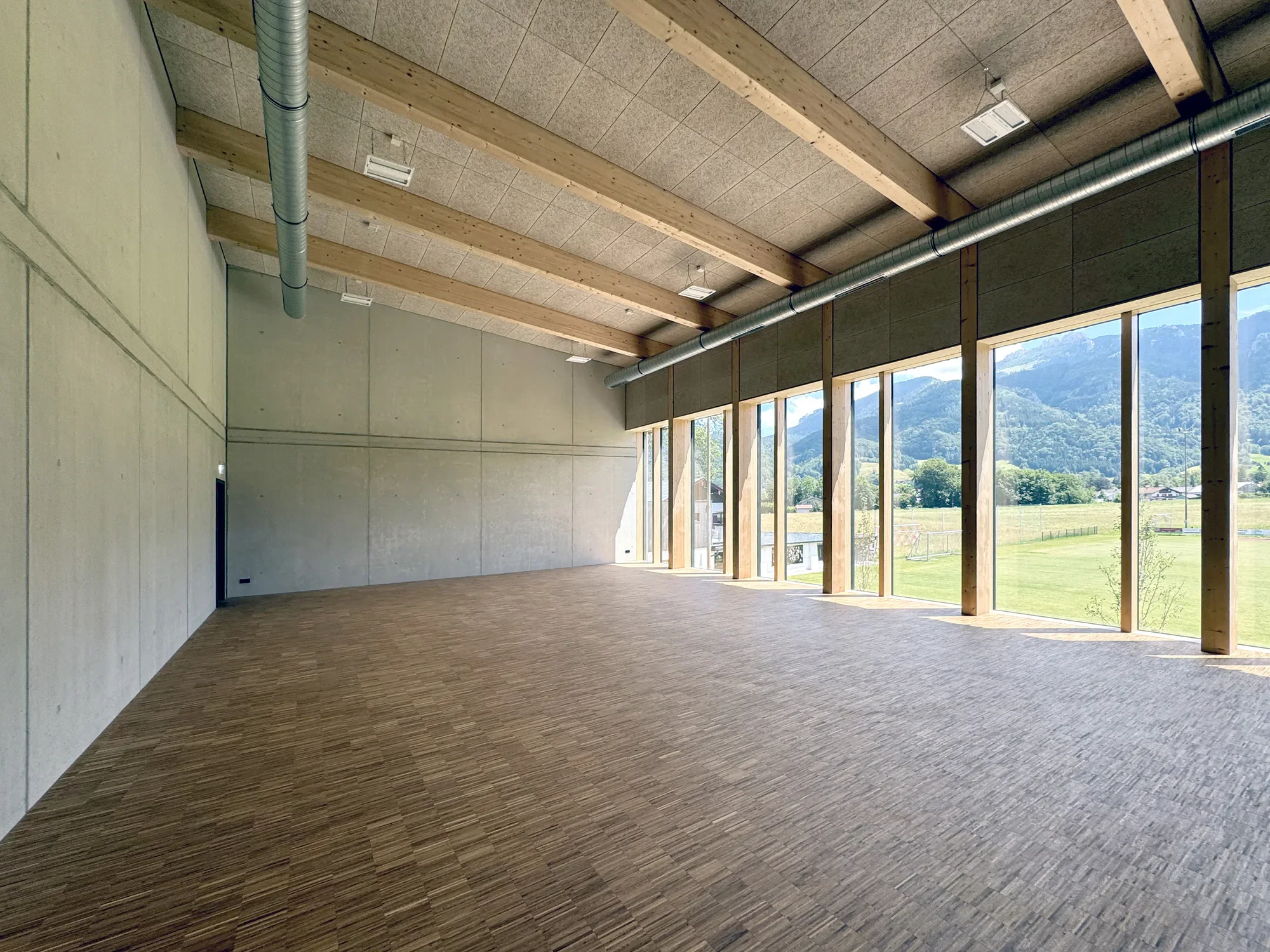 Ein großer, heller Innenraum mit Holzbalkendecken und großen Fenstern, die eine Berglandschaft mit grünen Wiesen und Bergen im Hintergrund zeigen.
