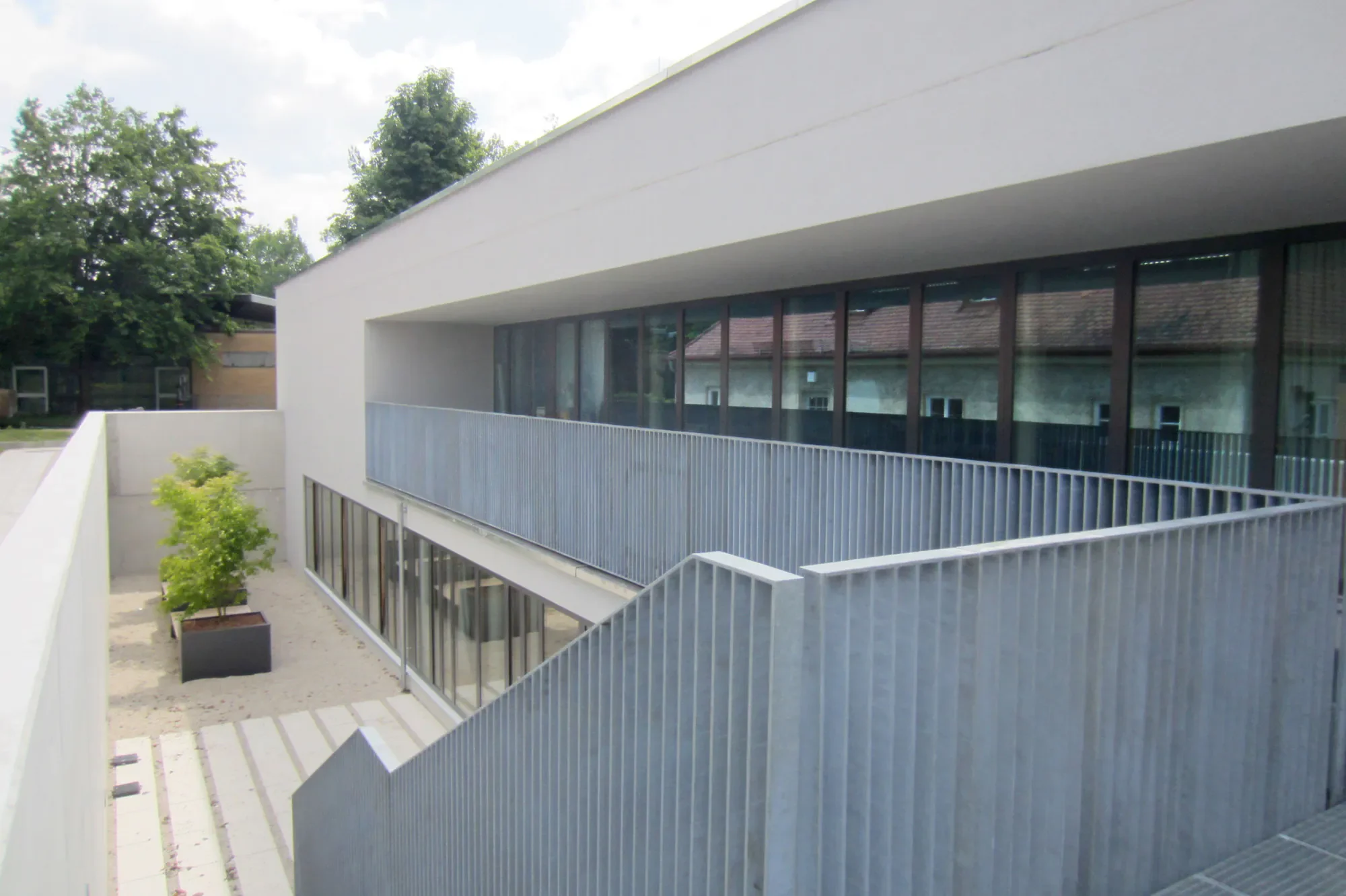 Moderne weiße Gebäudefassade mit großen Fenstern und Balkon, umgeben von grünen Bäumen, Blick auf einen Innenhof mit Weg und Pflanzkübel.