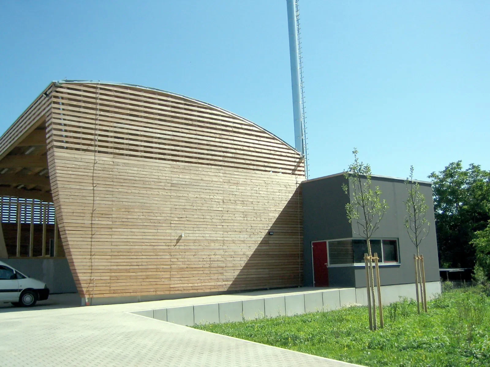 Moderne Gebäude mit Holzfassade und kleinen Bäumen im Vordergrund, blauer Himmel im Hintergrund.