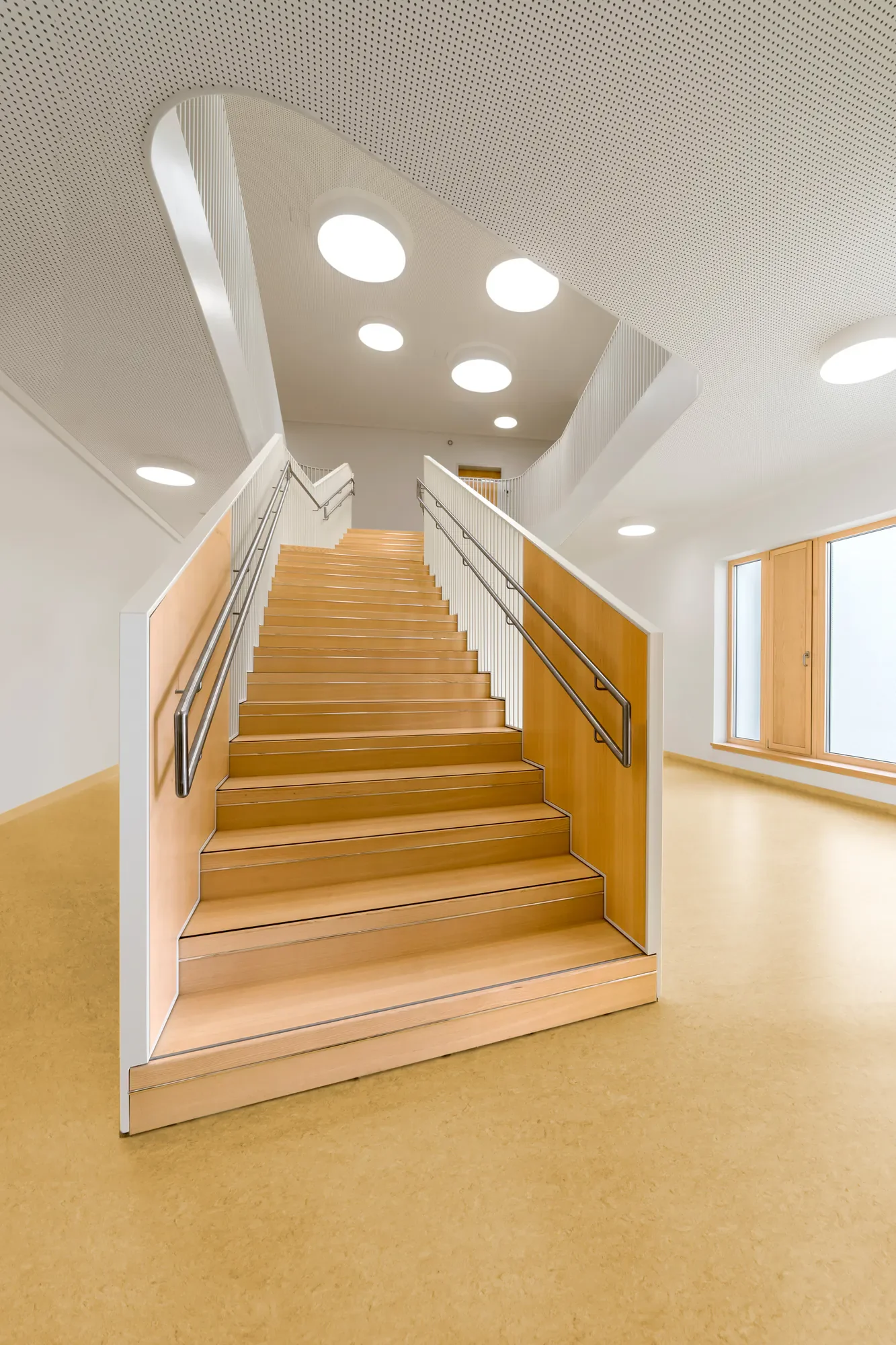 Innenansicht eines modernen Treppenhauses mit Holztreppe, weißen Geländern, großen Fenstern und runden Deckenleuchten.