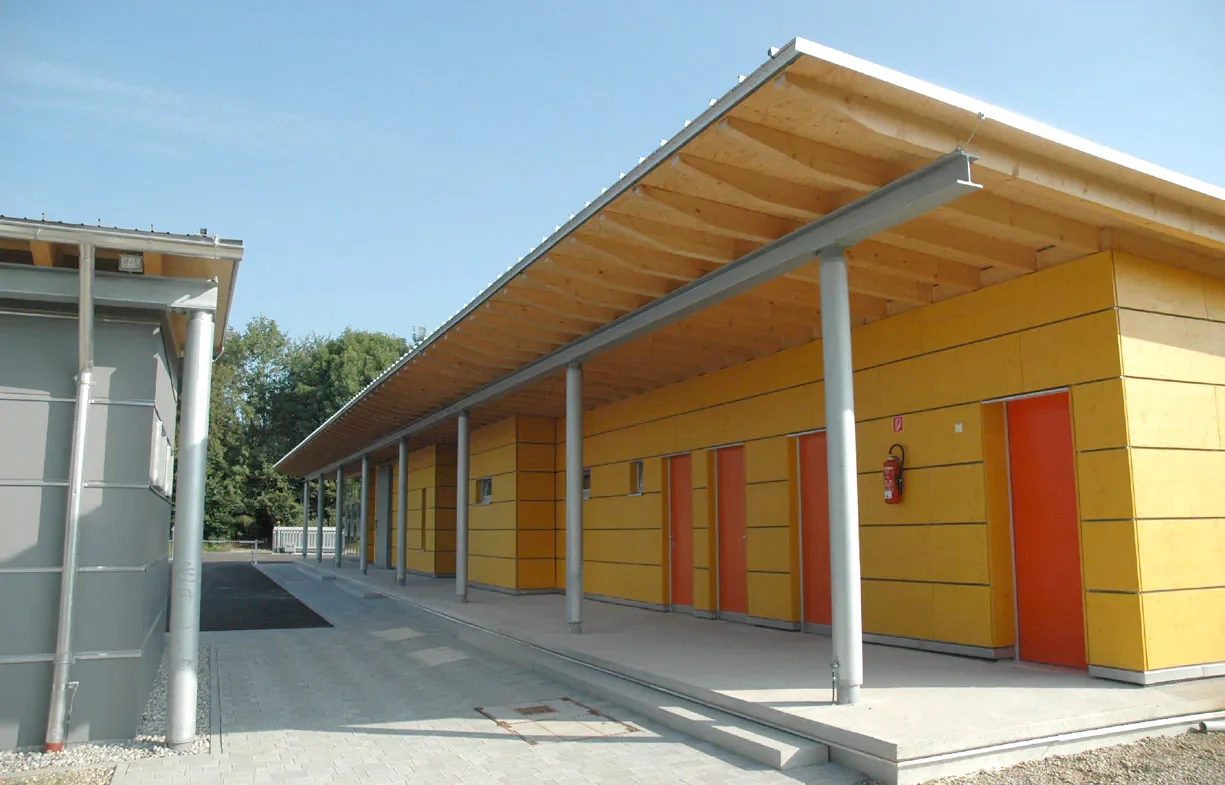 Ein modernes Gebäudekomplex mit gelben Holzfassaden und orange Türen, darüber eine große Überdachung mit Holz und Stahlstützen.