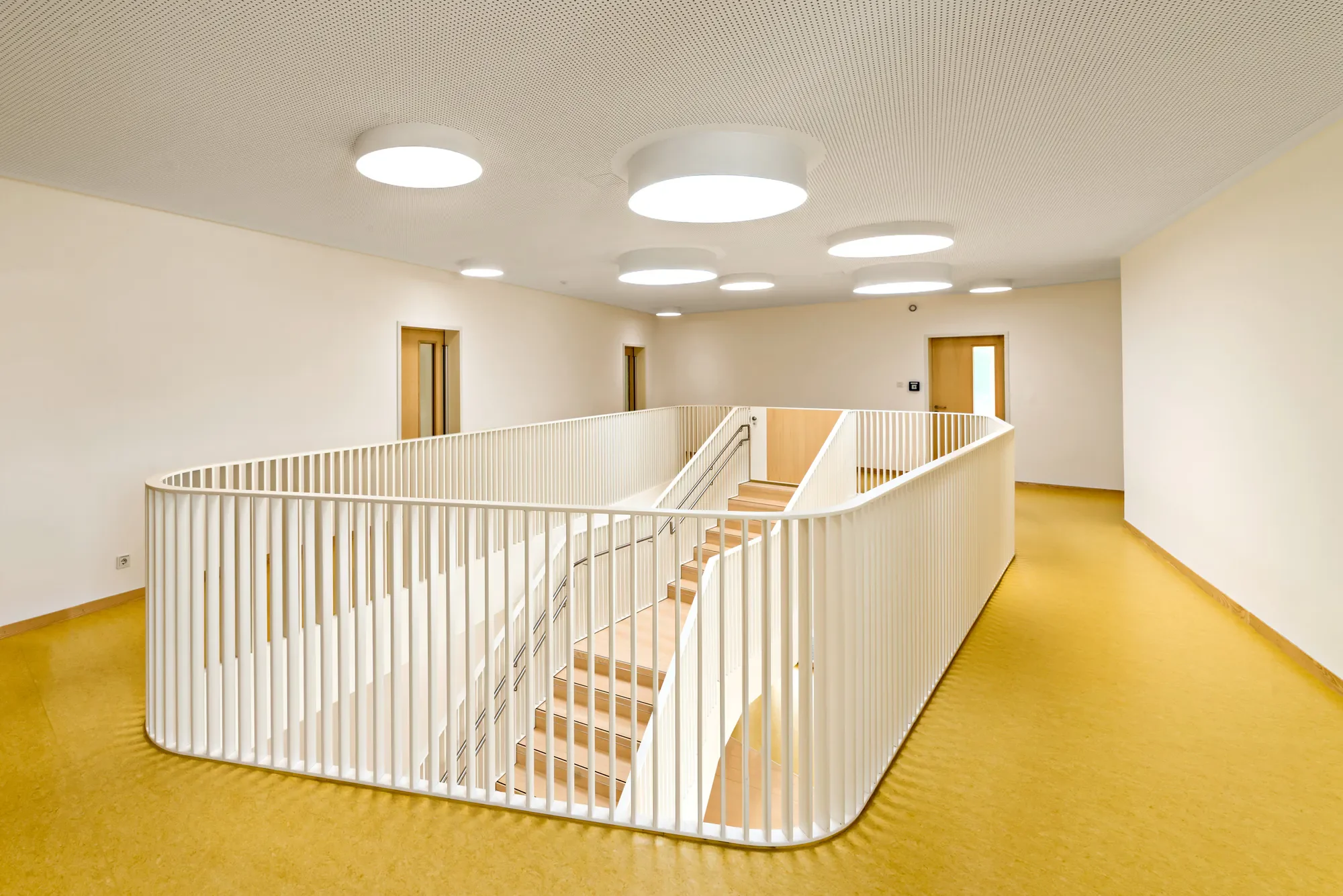 Innenansicht eines modernen Gebäudes mit weißer Treppe, gelbem Bodenbelag, weißen Wänden und runden, weißen Deckenleuchten.