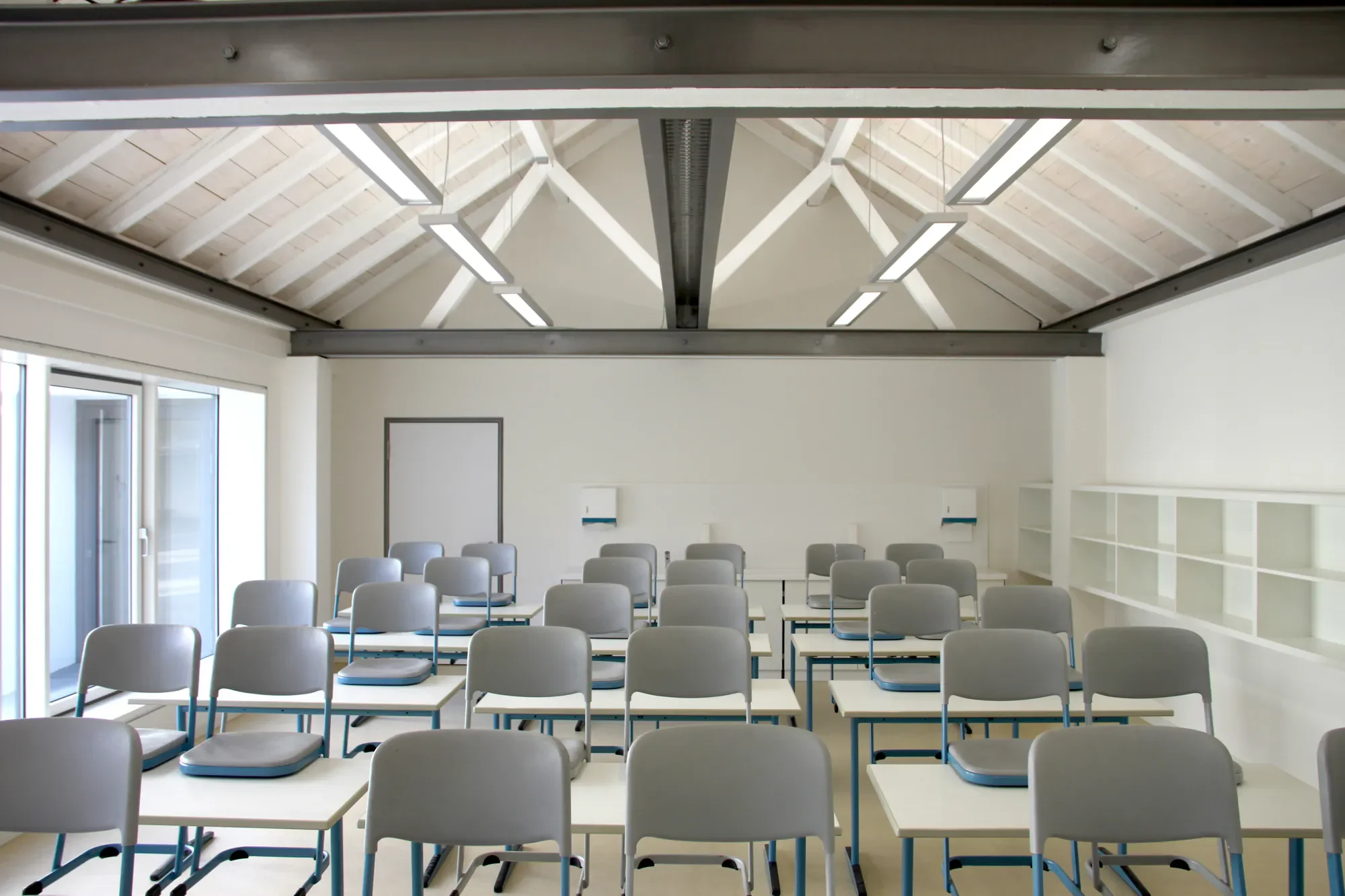 Ein heller, moderner Seminarraum mit weißen Stühlen und Schreibpulten, großen Fenstern und einer hellen Decke mit Holzträgern.