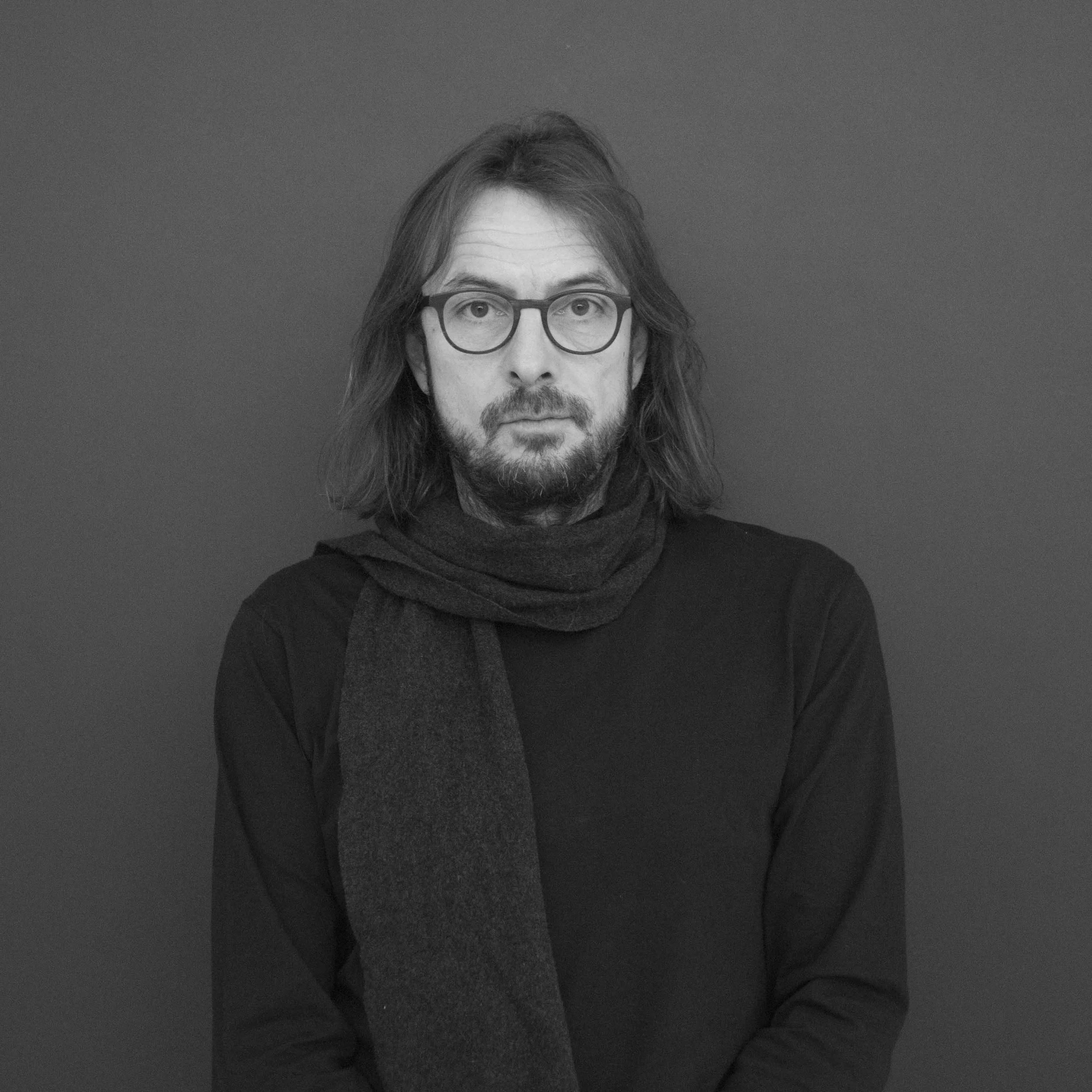 Schwarz-Weiß-Foto eines Mannes mit Brille, langem Haar und Bart, der einen schwarzen Pullover und einen Schal trägt, vor einem einfachen Hintergrund.