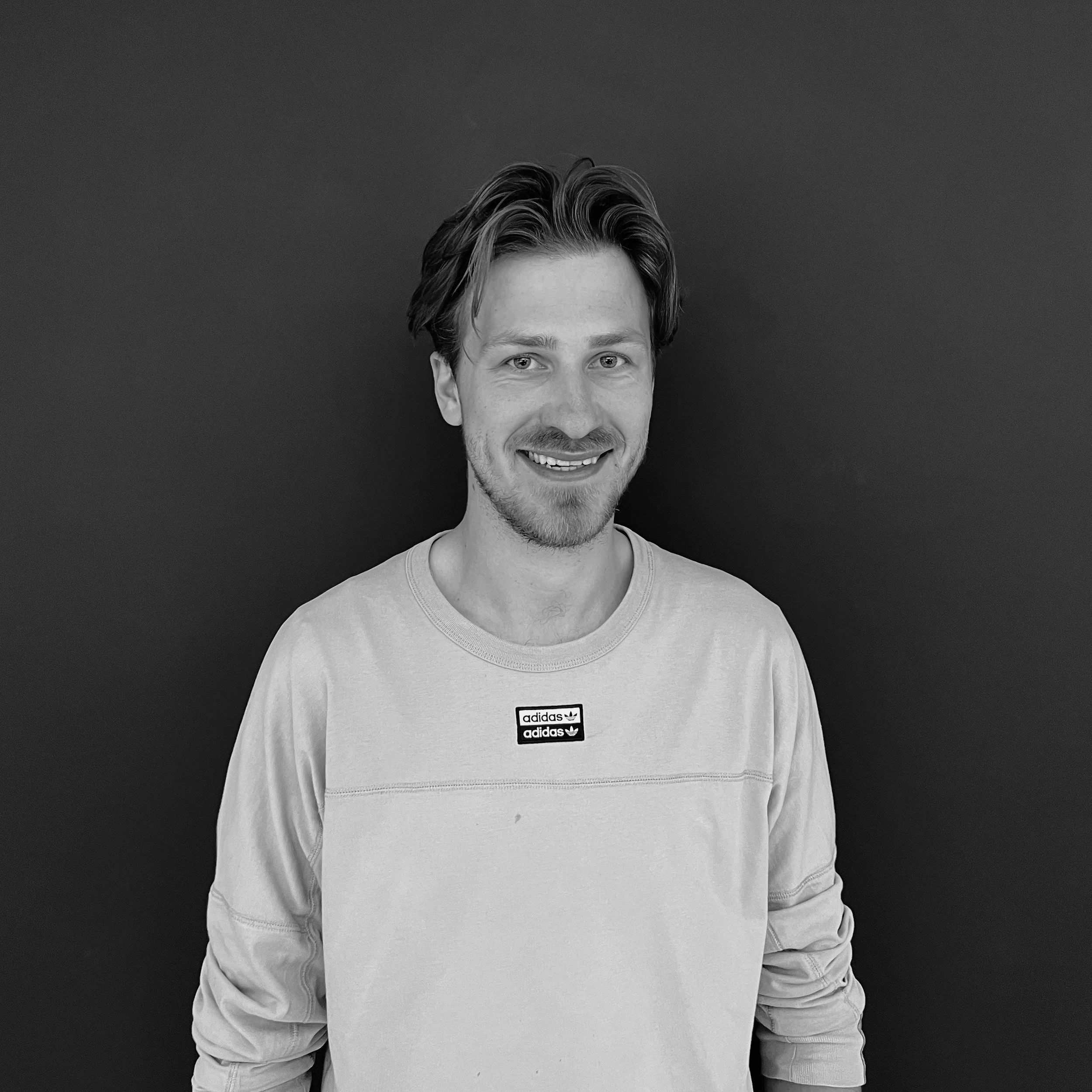Ein junger Mann mit braunen, gewellten Haaren, freundlichem Lächeln und einem grauen Adidas-Sweatshirt vor einem dunklen Hintergrund.