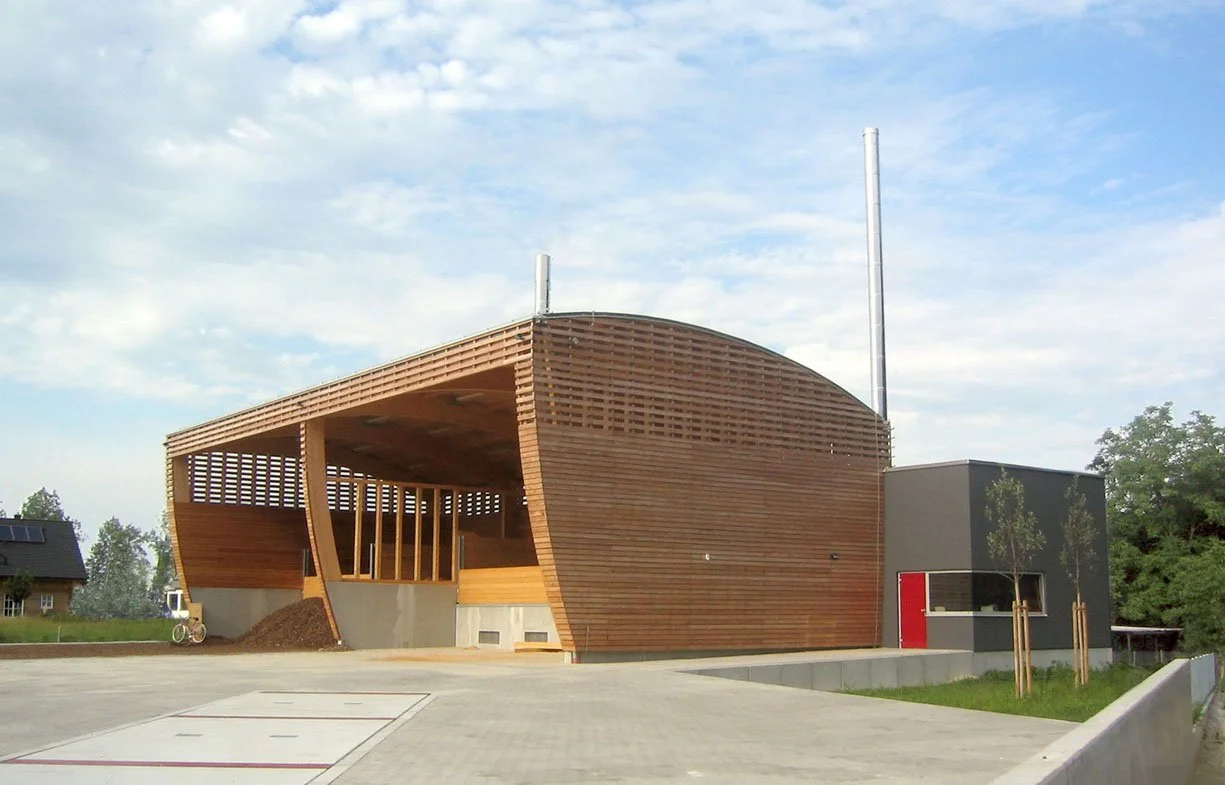 Modernes Gebäude mit einer einzigartigen, halbkuppelförmigen Holzfassade und grauen Betonfundament, umgeben von einem gepflasterten Bereich mit einigen jungen Bäumen.