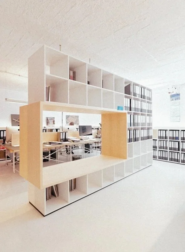 Offenes Büro mit weißen Regalen und mehreren Arbeitsplätzen mit Computern, einer großen Raumspartafel und hellem Ambiente.