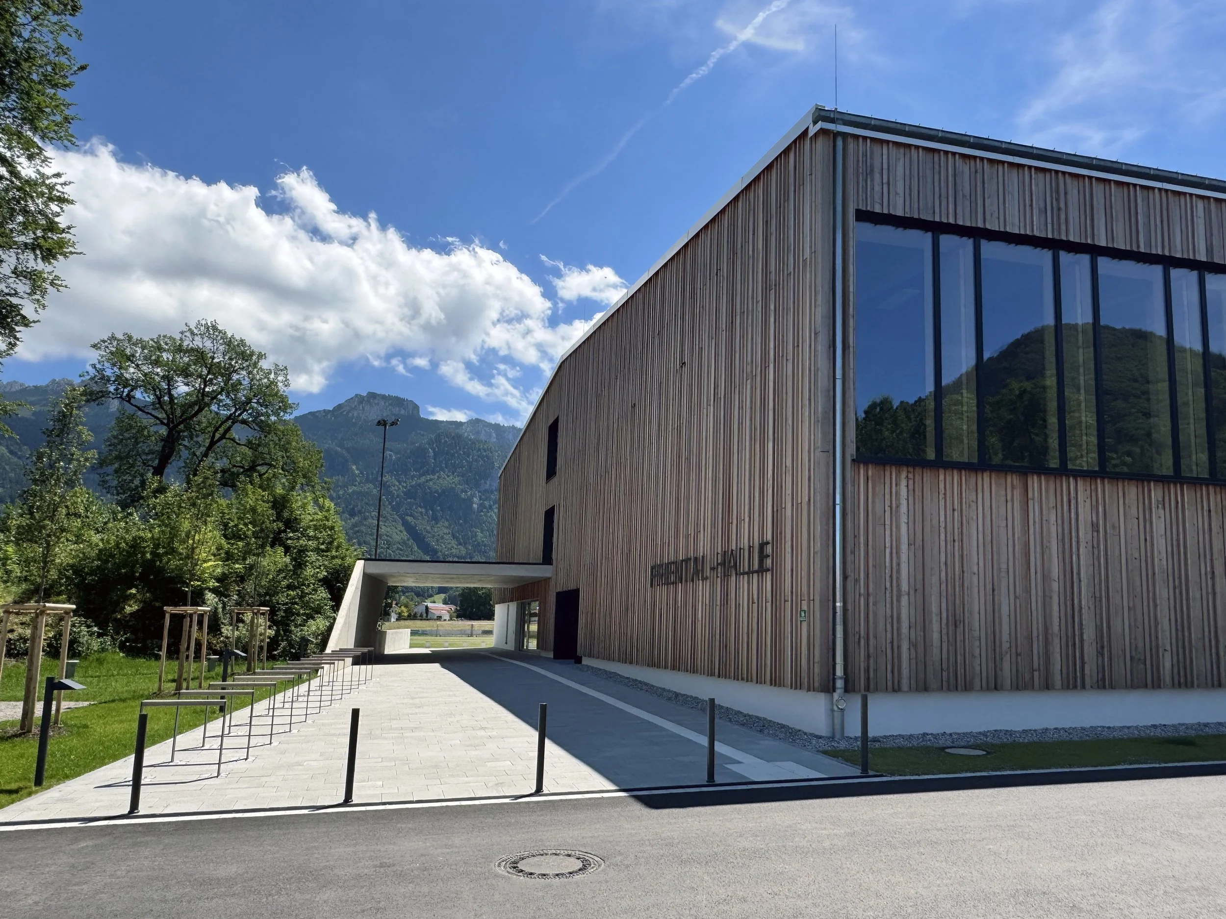Modernes Holzgebäude mit großen Fenstern, auf dem die Aufschrift «BEHANDLUNGSHALLE» steht, umgeben von grünen Bäumen und Bergen unter blauen Himmel mit Wolken.