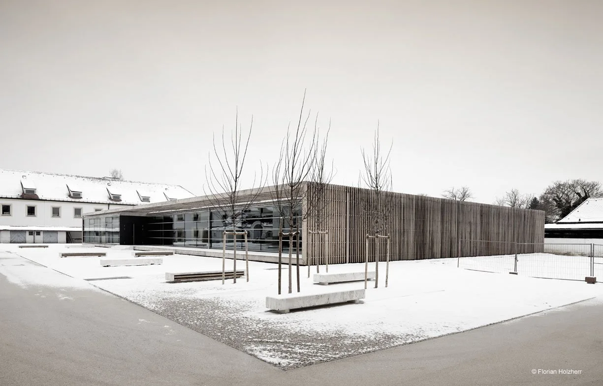 Ein modernes, flaches Gebäude mit Holz- und Glasfassade in einer schneebedeckten Umgebung, umgeben von jungen Bäumen und einer leeren, winterlichen Landschaft.