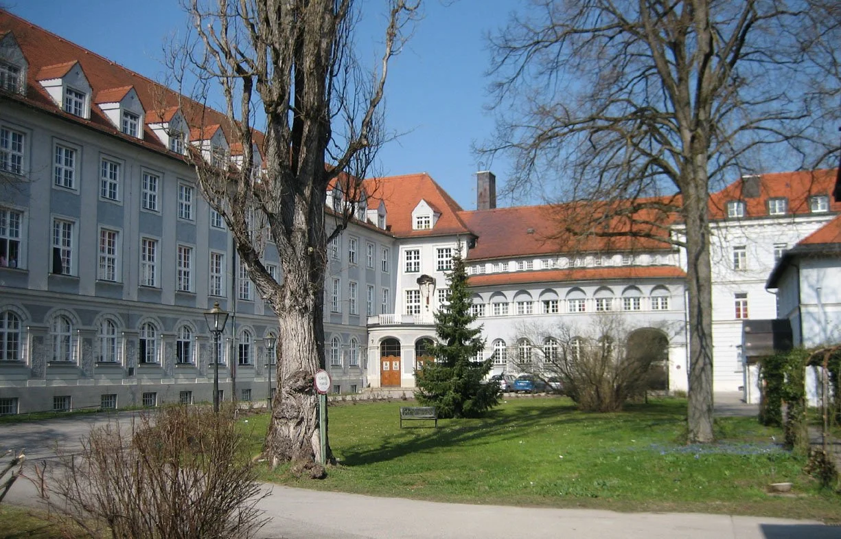 Ein Gebäudekomplex mit weißen Wänden und roten Dächern, umgeben von Bäumen und einem grünen Rasen, an einem sonnigen Tag.